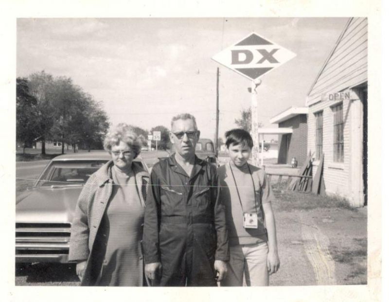 black and white photo of man and woman standing in front of a car at old gas station